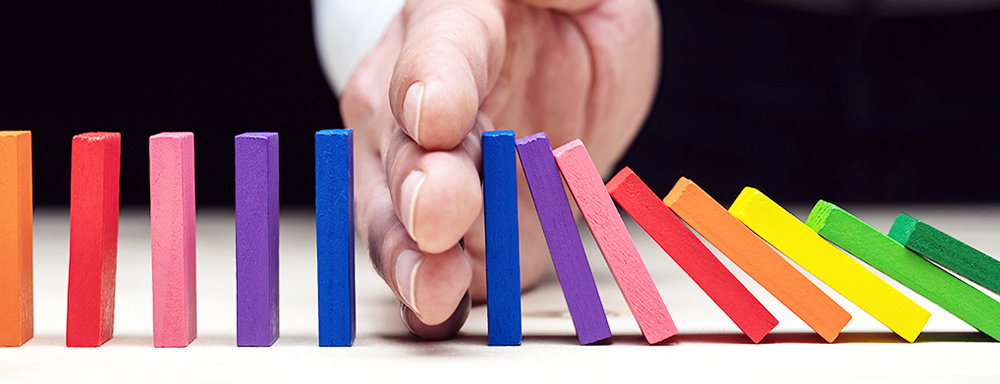 Eine Hand stoppt bunte Dominosteine, damit sie nicht weiter umfallen